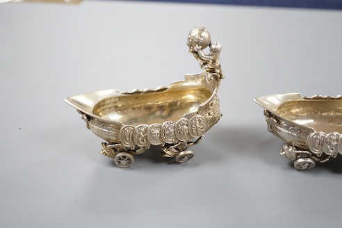 A pair of late 19th/early 20th century Hanau white metal salts, modelled as boat's with wheels and putti surmounts, maker Stork & Sinsheimer, length 81mm, 195 grams.